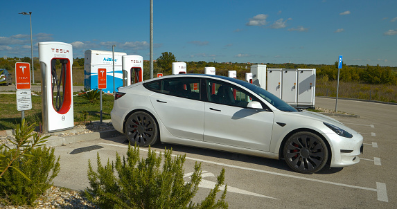Shell gas station in Croatia, 05.05.2023: CLOSE UP: Parked white electric car at Tesla charging point at battery refill. Plugged BEV car efficiently recharging empty lithium battery in the motor.