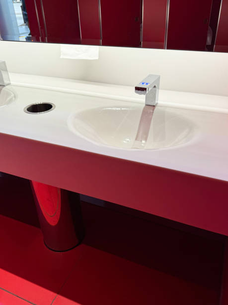 istock Image of row of inset sinks in white public restroom counter, rectangular bar, chrome, infrared basin mixer tap, mirrored wall reflecting row of red, toilet cubicle doors, focus on foreground 2077760442