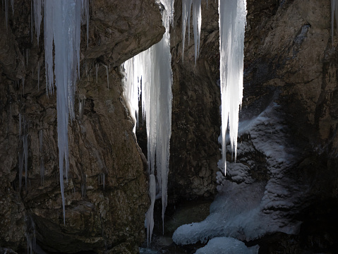 CLOSE UP: Glorious icicles hang above alpine stream flowing through narrow gorge. Backlit ice formations created by the freezing of trickling water. Amazing creations of nature in cold winter season.