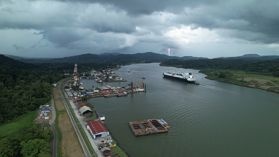Storm clouds rolling over port and cargo ship travelling through Panama Canal. Artificial water channel that runs through tropical environment and connects Atlantic and Pacific oceans on a stormy day.