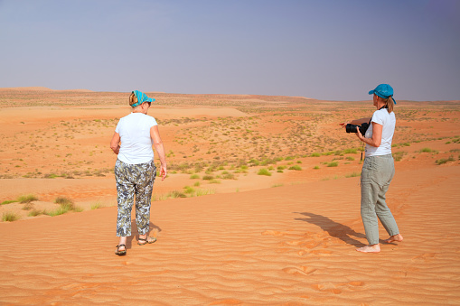 Back view of two senior woman, model and photographer on Wahiba desert sand in Oman. They are looking for memory photo with horizon, sand and sky.