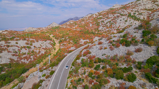 AERIAL: Stunning autumn coastal landscape and car driving along the asphalt road. Panoramic highway in the embrace of rugged Mediterranean terrain with shrubs glowing in vivid shades of fall season.