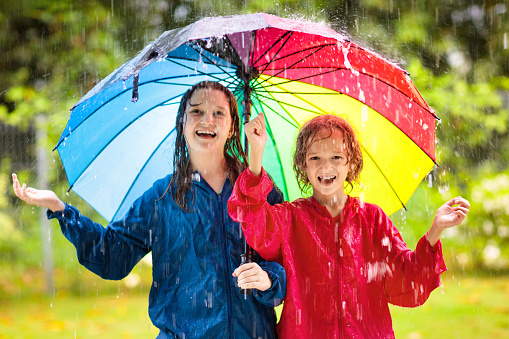 Kids play in heavy autumn rain. Children walk on city street in cold rainy weather. Waterproof warm outdoor wear. Boy and girl with colorful umbrella and rain jackets. Outdoors fun.