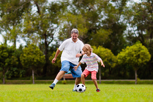 Father and son play football on outdoor pitch. Dad and kid run on park stadium. Man and little boy playing soccer on sunny summer day. Healthy active sport for young family.