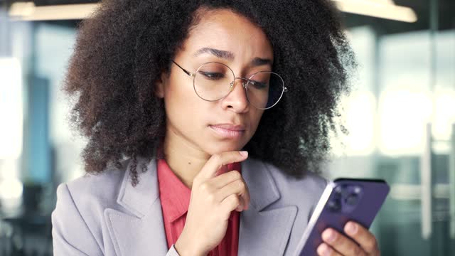 Thoughtful serious young african american businesswoman is using phone sitting at workplace in business office.