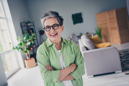 Photo of cheerful senior funny woman alone but friendly businesswoman folded arms wear glasses laughing isolated indoors apartment room.