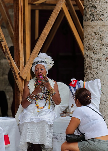 Havana, Cuba-October 7, 2019: Cuban santera with spectacular nails serves a client requesting her divination services while smoking a big cigar under the arcade of the Plaza Catedral Square east side.