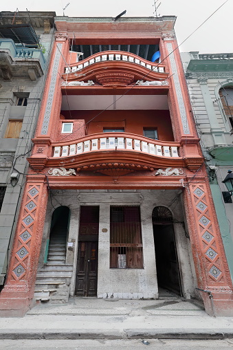 Dilapidated facade of three-story tenement house in Centro Havana with undulating balconies and imposing portal topped by dragon reliefs, some steps missing from the entrance stairway. La Habana-Cuba.