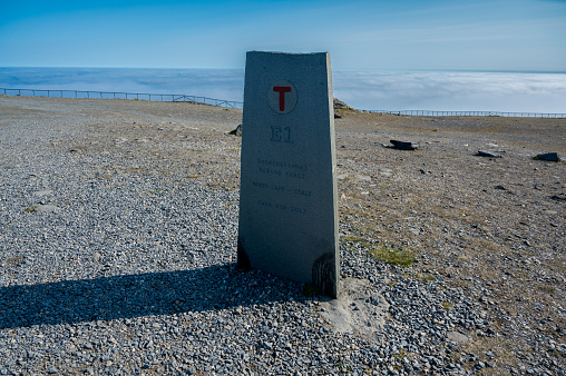 Monument to the international hiking trail E1 in Nordkapp, Norway, with clouds under cliffs