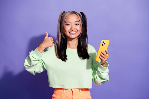 Portrait of millennial girl wear sweatshirt thumb up symbol like feedback for new iphone 15 features isolated on purple color background.