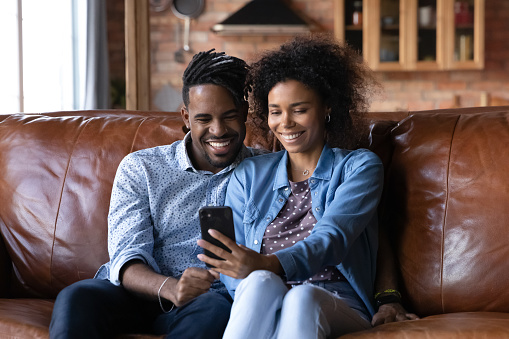 Attractive African couple sit on sofa looking at cellphone screen, make video call use mobile app, watch internet content, taking selfie pictures, spend leisure at home enjoy modern tech usage concept
