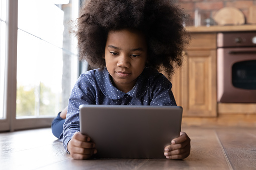 Little 6s African girl lying on warm floor with digital tablet, look at device screen watch online cartoons, reading e book, close up. Modern technology overuse, parental control app, safety concept