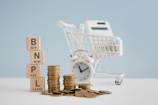 For buy now pay later , online shopping concept, wooden cube block with BNPL text and stack of coins, blurred white calculator in shopping cart and clock