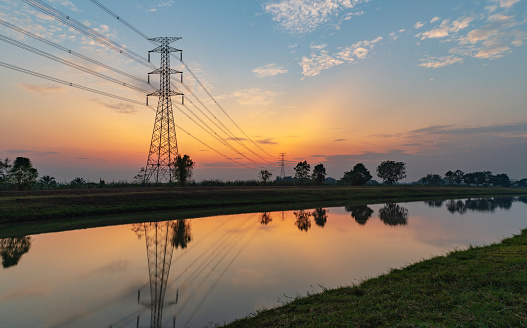 The silhouette of a high-voltage electric pole in the countryside alongside a water canal.