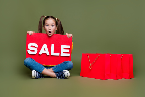 Full length photo of speechless kid with ponytails hairdo sit on floor hold sale poster staring at sale isolated on khaki color background.
