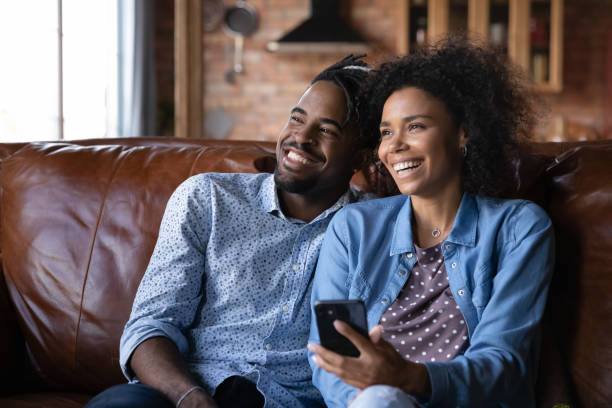 Cheerful African couple sit on sofa at home with cellphone