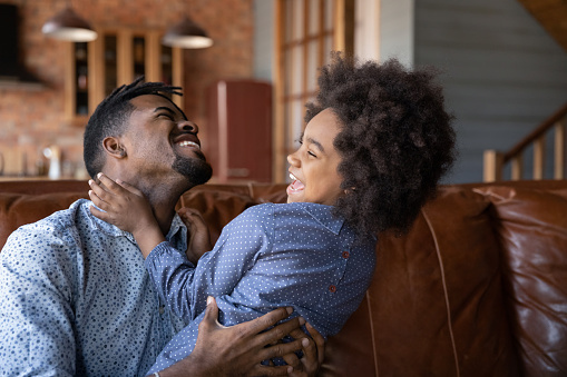 Young African dad having fun with little cute 6s curly-haired daughter, cheery family sit on sofa laughing tickling each other enjoy carefree playtime at home. Happy fatherhood day, love, bond concept