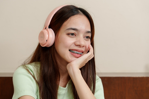 Young Asian woman with dark hair, wearing pale green top, enjoys music on pink headphones, hint of joy in smile. Music enthusiast in light-hued sweater lost in melody, pink over-ear audio gear