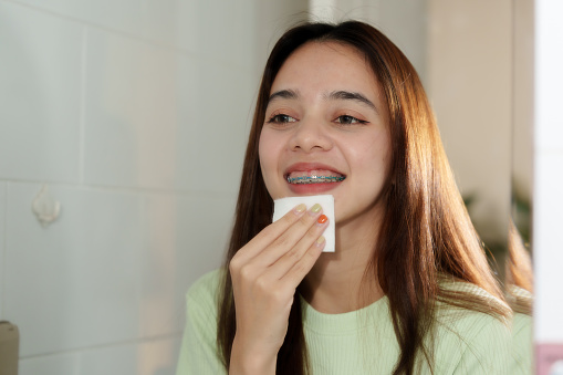 Asian Young adult with long hair smiles while holding white card to forehead, skincare routine, in tiled bathroom. braces exhibits skincare regimen using cotton pad in well-lit, displaying self-care.