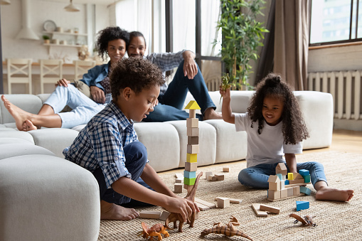 Preschool African siblings play wooden cubes and dino toys sit on warm floor in living room, their parents relaxing on sofa. Family weekend leisure at home, modern playthings, kids development concept
