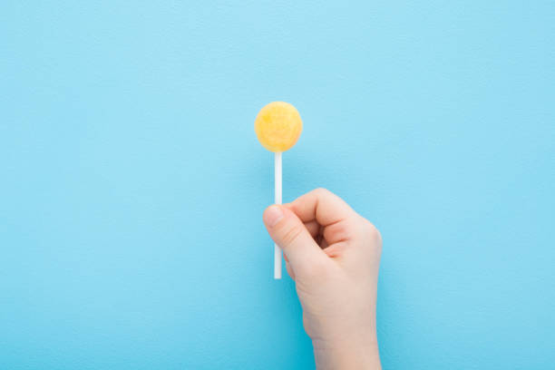 Little child fingers holding yellow lollipop on stick on light blue table background. Pastel color. Sweet candy. Closeup. Top down view.