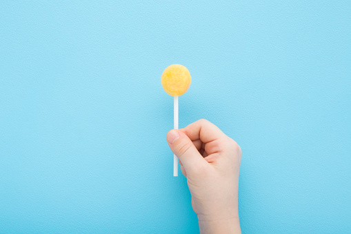 Little child fingers holding yellow lollipop on stick on light blue table background. Pastel color. Sweet candy. Closeup. Top down view.