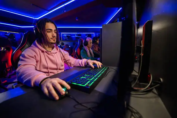 Young male cybersport gamer with dreadlocks and a mustache sitting in front of a computer monitor in cyber club and playing a video game with his team. Esport streaming game online. Copy space.
