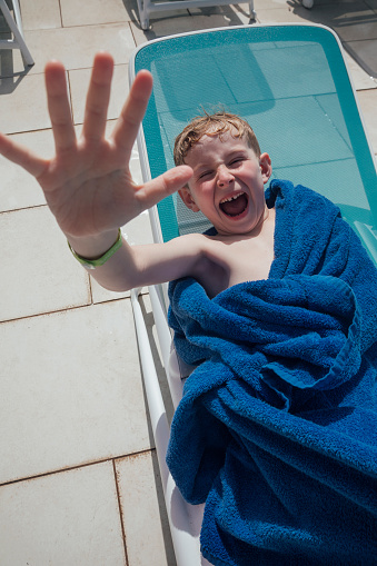 A young boy wearing a towel, lying down on a sun lounger at an all inclusive hotel in Tenerife, Spain. He has his arm raised to the camera while looking at it with an excited look on his face.