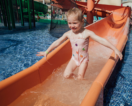 A young girl wearing a swimsuit on holiday, playing at a waterpark at an all inclusive hotel in Tenerife, Spain. She has just slid down a slide and is standing in the water at the bottom of the slide while holding onto it and looking forward with a smile on her face.