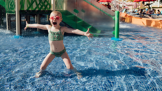 A young girl wearing a bikini and goggles on holiday, playing at a waterpark at an all inclusive hotel in Tenerife, Spain. She is standing in a swimming pool with her arms outstretched while looking at the camera with her mouth open excitedly.