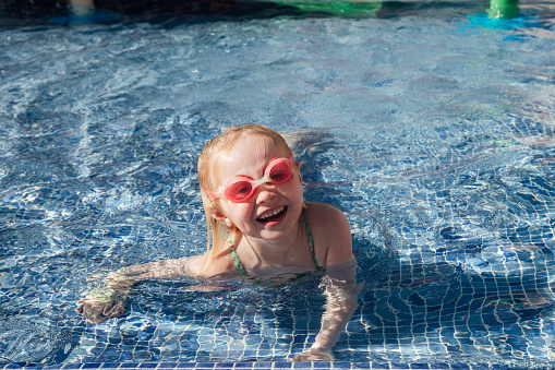 A young girl wearing a swimsuit and goggles on holiday, playing in a swimming pool at an all inclusive hotel in Tenerife, Spain. She is lying in the water while looking at the camera and smiling.