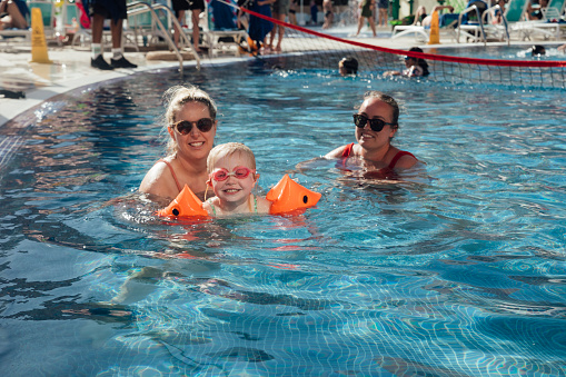 A young girl wearing armbands and goggles, swimming with her two aunties in a swimming pool at an all inclusive hotel in Tenerife, Spain. They are all looking at the camera and smiling.