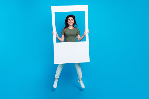 Full size photo of gorgeous positive cute woman wear green t-shirt white trousers jumping hold frame isolated on blue color background.