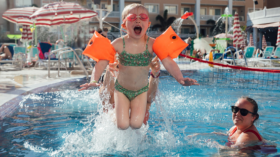 A young girl wearing armbands and goggles, being thrown into a swimming pool by her auntie who is behind her in the pool at an all inclusive hotel in Tenerife, Spain. Her other auntie is looking at her and smiling as she descends into the water.