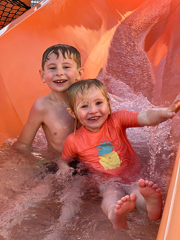 Two children wearing swimwear, sliding down a waterslide at a waterpark at an all inclusive hotel in Tenerife, Spain. They are looking at the camera and smiling as they head down the slide.