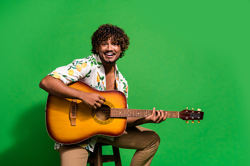 Photo portrait of attractive young man sit chair play guitar dressed stylish citrus print clothes isolated on green color background.