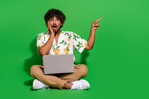 Full body photo of man sit floor shocked point empty space netbook dressed stylish citrus print clothes isolated on green color background.