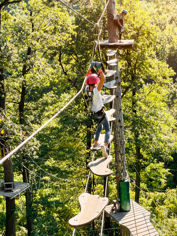 Rope park in wood forest.Adventure park close up. Summer fun and sports for adventurous people.Happy A teenager Girl enjoying activity in a climbing adventure park walks along a rope bridge between trees in safety gear and a helmet on a summer day.