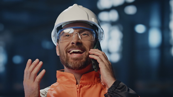 Joyful factory worker calling at manufacturing facility close up. Portrait happy engineer in protective helmet uniform talking cellphone feeling excited. Smiling workman friendly phone conversation