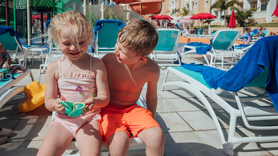 Two children wearing swimwear, sitting on a sun lounger at an all inclusive hotel in Tenerife, Spain. They are smiling as the sun beats down on them and the young boy is looking at his sister in awe.