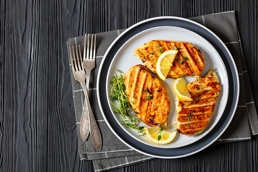 boneless skinless juicy grilled chicken cutlets with lemon slices on plate on black wooden table with forks, horizontal view from above, flat lay, free space