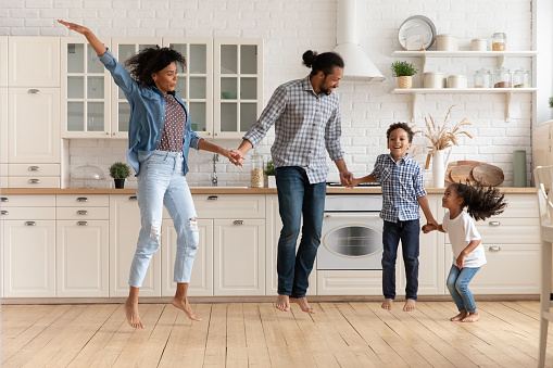 Beautiful African couple and little cute children holding hands jumping barefoot together in fashionable kitchen on warm wooden floor. Active weekend, new modern house, happy homeowners family concept