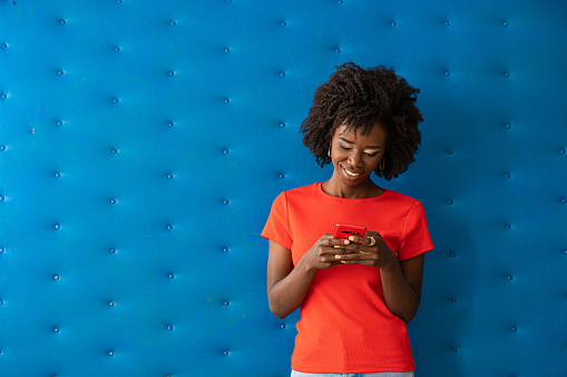 Studio shot of an attractive african young woman, wearing red t-shirt, posing in front of blue background and talking on a smart phone