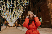 Portrait of amazing redhead young woman in hat and winter jacket standing posing on snow city street on blurred background of hinged bright festive illumination.