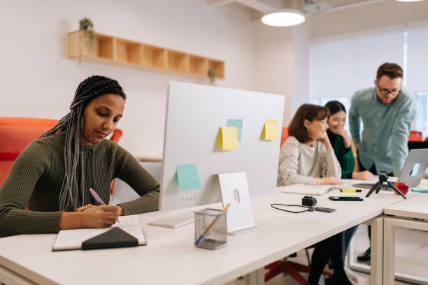 istock Focused African-American businesswoman writing down notes and using computer sitting at work in big modern corporate office. Colleagues businesspeople working together 2078313952