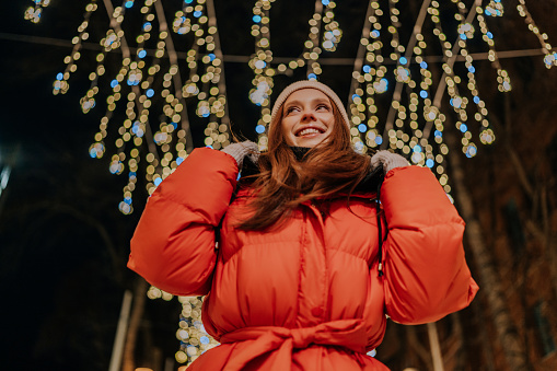 Low-angle view of happy young woman in warm hat and winter jacket standing posing on snow city street on blurred background of hinged festive light illumination. Pretty female walking on square.