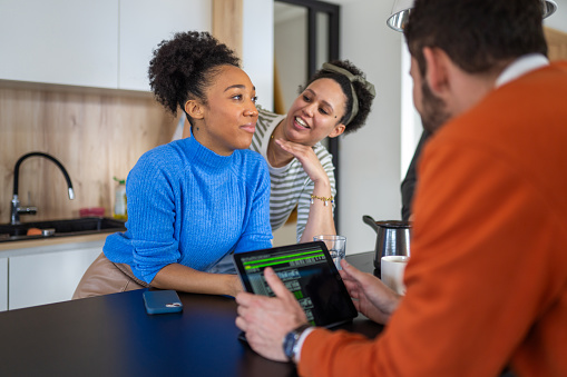 Two female and one male young adults in casual attire engaged in a dynamic conversation about investment strategies at a modern kitchen table. A digital tablet displaying financial graphs is in use, indicating a focus on market trends and data analysis.