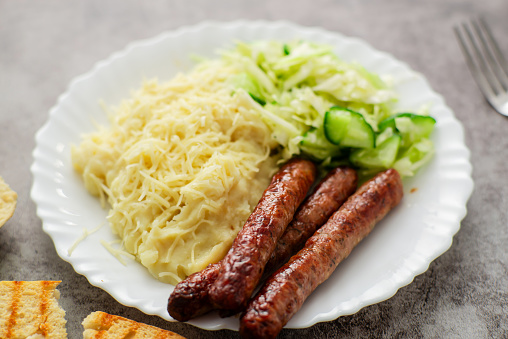 Indulge in a comforting meal of cheese-infused mashed potatoes, a crisp cabbage and cucumber salad, and flavorful sausages, beautifully arranged on a white plate against a dark backdrop