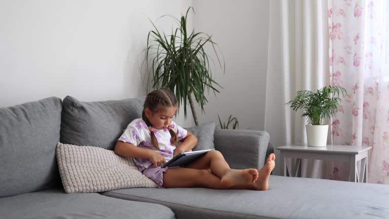 Adorable little girl sits at home on sofa and play games on tablet. Home mood