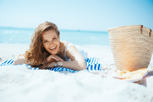 smiling modern middle aged woman on the ocean coast with straw bag and striped towel.